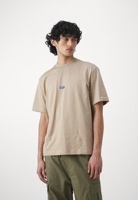 Beige bomulds t-shirt med kortærmer og rund halsudskæring, med et lille blåt og hvidt logo på brystet. Glat tekstur.