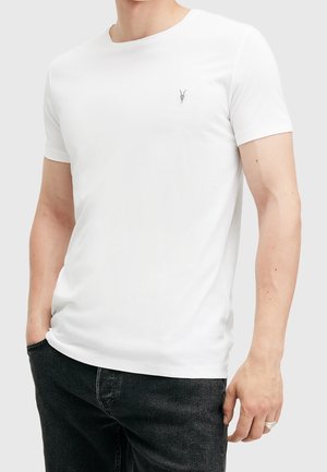 Homme portant un t-shirt blanc uni à manches courtes et col rond avec un petit logo tête de chèvre noir, associé à un jean noir.