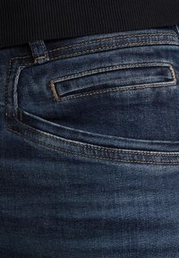 Jeans iz denima v temno modri barvi, z zadnjim žepom s podrobnostmi šivanja in gladko teksturo, delno pokriti s črnim vrhom.