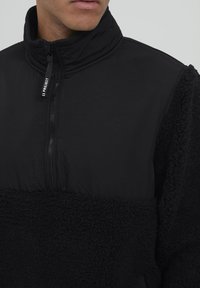 Schwarzer Fleece-Pullover mit hohem Kragen und Reißverschluss. Die obere Hälfte besteht aus glattem Stoff, während die untere Hälfte strukturiert und flauschig ist.