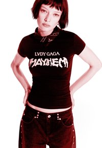 Mladý člověk s krátkými tmavými vlasy, oblečený v černém tričku "Lady Gaga Mayhem" a tmavých kalhotách, s rukama opřenýma o boky, na světlém pozadí.