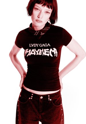 Giovane con capelli corti e scuri, che indossa una maglietta nera "Lady Gaga Mayhem" e pantaloni scuri, con le mani sui fianchi, su uno sfondo chiaro.