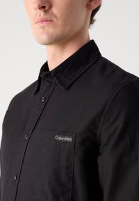 Črna srajca z gumbi iz bombažnega materiala z nazobčanim ovratnikom in žepom na prsih. Ima belo oznako z logotipom "Calvin Klein" na žepu.