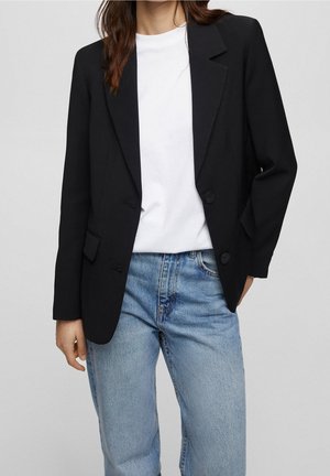 Femme portant un blazer noir par-dessus un t-shirt blanc et un jean taille haute bleu clair, debout avec une main dans la poche sur un fond gris.