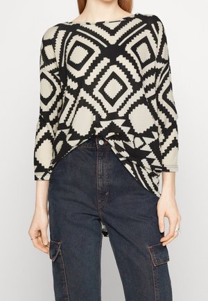 Woman wearing a black and beige geometric patterned long-sleeve top tucked into dark blue high-waisted cargo jeans with side pockets.