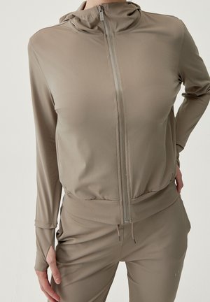 Person iført en taupe lynlåshoodie med tommelfingerhuller og matchende joggingbukser, stående mod en ensfarvet lys baggrund.