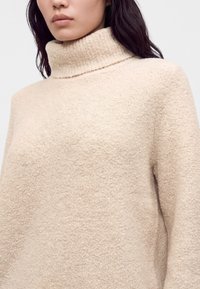 Femme porte un pull à col roulé texturé couleur crème à manches longues, montré de la poitrine vers le haut sur un fond uni.