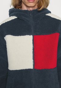 Fleece zip-up jakke med hætte, der har navy, creme og røde paneler; tekstureret, blødt stof; frontlynlås og afslappet design.