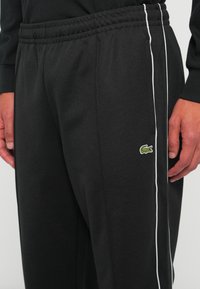 Zwarte trainingsbroek met elastische tailleband, witte zijkanten en een klein groen krokodillenlogo op de rechterdij, gedragen door een persoon.