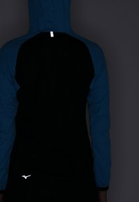 Hooded jacket featuring a black body with blue sleeves, smooth texture, and reflective logo detail near the hem. Back view shown.