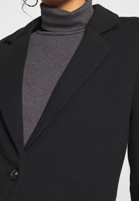 Sort blazer med almindelig krave, enkelt knaplukning og ribbet mørkegrå turtleneck underneath. Glat stoftekstur.