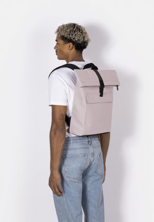 Man with curly hair wearing a white t-shirt and jeans carries a light pink rectangular backpack with black straps.