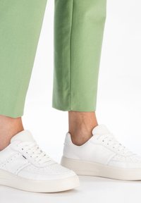 Weiße Leder-Sneaker mit durchbrochenen Details, flachen Schnürsenkeln und strukturierten Sohlen, kombiniert mit grünen, knöchellangen Hosen.