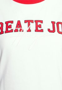 Hvid t-shirt med rød halsudskæring. Har fed "CREATE JO" tekst med sort omrids og en subtil signatur i lys pink.