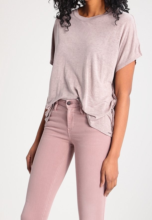 Kobieta ubrana w luźną, jasnofioletową koszulkę, lekko włożoną do różowych, dopasowanych dżinsów z wysokim stanem, stojąca z jedną ręką swobodnie opuszczoną wzdłuż ciała.