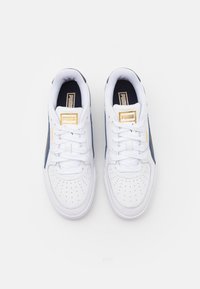 Paire de baskets blanches Puma avec côtés bleus, logo Puma doré sur la languette et le côté, vue de dessus sur fond blanc.