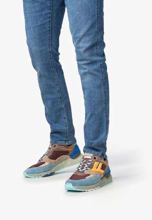 Person, die blaue Jeans und mehrfarbige Sneaker mit blauen, braunen, beigen und orangen Details trägt, steht vor einem weißen Hintergrund.