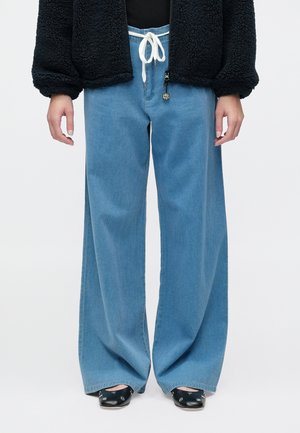 Personne portant un jean ample large bleu clair avec un cordon blanc, une veste noire texturée et des ballerines noires à clous, debout les bras détendus.