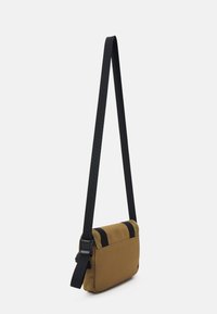 Braune Canvas-Umhängetasche mit schwarzem verstellbarem Tragegurt; zeichnet sich durch ein minimalistisches Design und zwei schwarze Akzentriemen an der Oberseite aus.