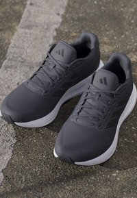 Paire de chaussures de course Adidas gris foncé avec semelles blanches sur l'asphalte à côté d'une ligne blanche délavée.