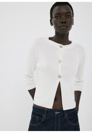 Persona de piel oscura que lleva un cárdigan corto blanco con tres botones decorativos y vaqueros de mezclilla azul oscuro contra un fondo liso.