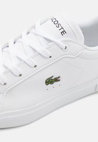 Valged nahast tossud valgete paeltega, millel on roheline krokodilli logo ja aukudega detailid küljel. Lamenev kummist tall.
