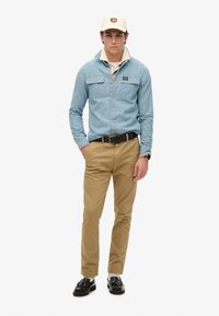 Lichtblauw denimshirt met twee borstzakken, beige chinos, zwarte riem en schoenen, gecombineerd met een lichtgekleurde pet en een witte onderhemd.