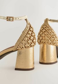 Sandales dorées à talon bloc, dos tissé et brides de cheville avec boucles, vues de dos sur un fond uni.