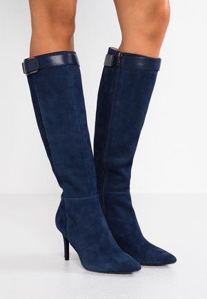 Marineblå ruskindsknæhøje støvler med spidse snuder, slanke hæle, sidelommer med lynlåse og teksturerede stropper med spænder øverst.