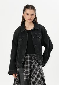 Giacca di jeans nera con una vestibilità rilassata, chiusura con bottoni, due tasche sul petto e maniche leggermente oversize. Indossata sopra a una maglietta nera con una camicia a quadri legata in vita.