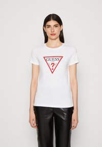 T-shirt bianco in cotone con un logo a triangolo rosso con la scritta "GUESS?" e ulteriore testo. Il modello indossa pantaloni in pelle nera.