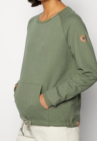 Sudadera de algodón verde con cuello redondo, bolsillos frontales, cordón ajustable en el dobladillo y un pequeño acento de cuero en la manga.
