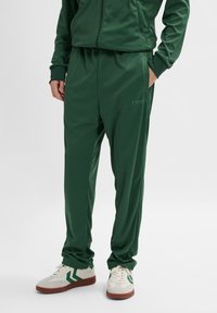 Grønne polyester træningsbukser med en elastisk talje, sidelommer og et lille logo-print. Båret med hvide sneakers med grønne accenter.