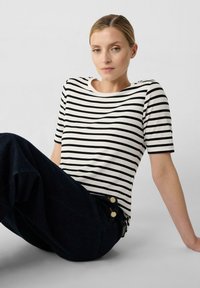 Gestreiftes kurzärmliges Oberteil in Creme mit schwarzen horizontalen Streifen. Es hat einen runden Halsausschnitt und eine lockere Passform, kombiniert mit dunklen Jeans.