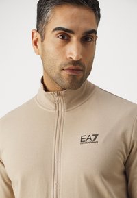 Bežna zip-up jakna iz raztegnjenega tkanine z visokim ovratnikom. Značilna je črna EA7 logo na levi strani. Gladka tekstura.