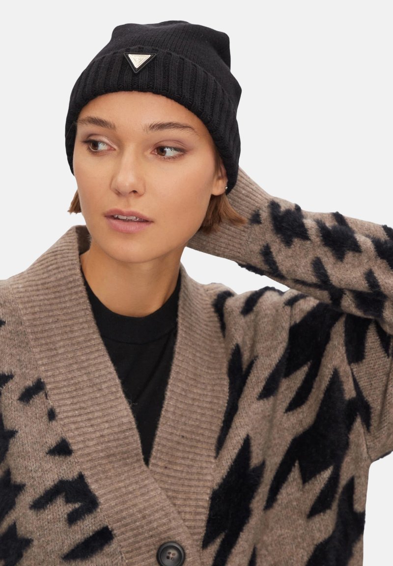 Cappello a cuffia nero a coste con un logo triangolare argento. Il modello indossa un cardigan beige con motivo e accenti scuri e una maglietta nera sotto.