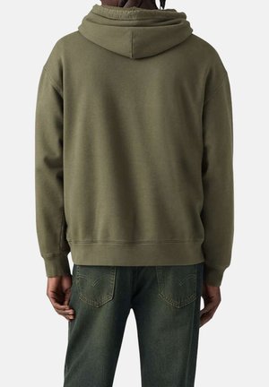 Personne portant un sweat à capuche vert olive et un jean vert foncé, vue de dos avec les mains détendues le long du corps.