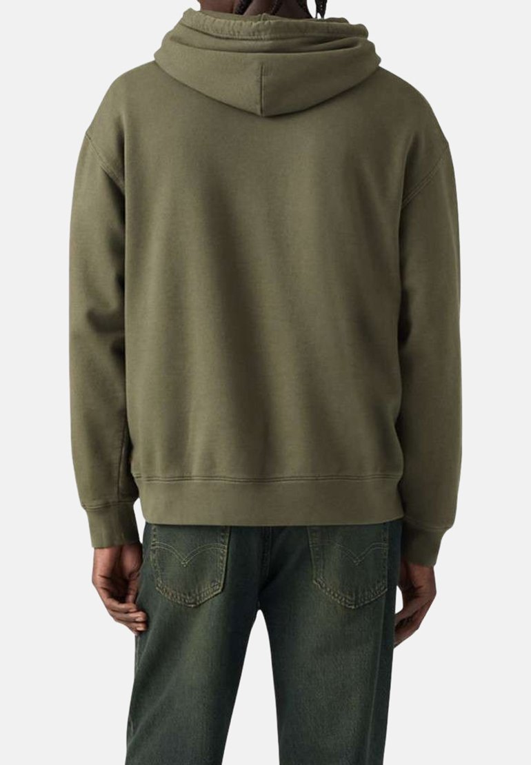 Persona vistiendo una sudadera con capucha verde oliva y jeans verde oscuro, de espaldas con las manos relajadas a los lados.