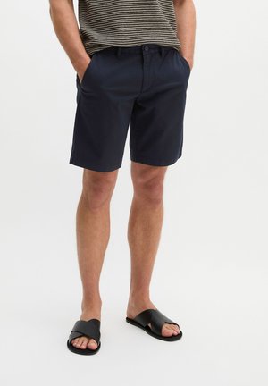 Mann trägt marineblaue Shorts, ein gestreiftes Hemd und schwarze Slip-On-Sandalen und steht mit den Händen in den Taschen vor weißem Hintergrund.