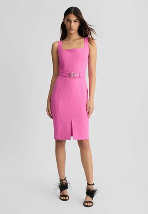 Robe ajustée rose avec décolleté carré, taille ceinturée ornée d'une boucle circulaire et fente à l'avant. Porte avec des sandales noires ornées de plumes.