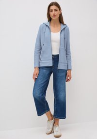 Felpa azzurra con zip e coulisse, indossata sopra una canotta bianca, abbinata a jeans blu a gamba larga e sneakers beige.