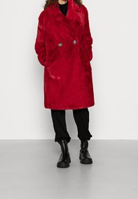 Punainen tekoturkistakki, jossa on kaksirivinen napitus, kaulukset, edessä taskut ja kiiltävät mustat napit, puettu mustien housujen ja nilkkureiden päälle.