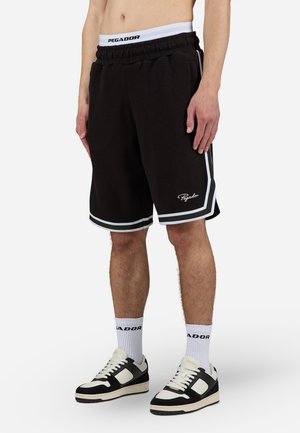 Mężczyzna w czarnych szortach do koszykówki Pegador z białymi lamówkami, białe skarpetki z logo Pegador oraz czarno-białe sneakersy stoi na jednolitym tle.