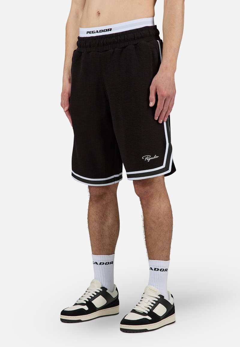 Homme portant un short de basketball Pegador noir avec des bordures blanches, des chaussettes blanches à la marque Pegador et des baskets noires et blanches, debout devant un fond uni.