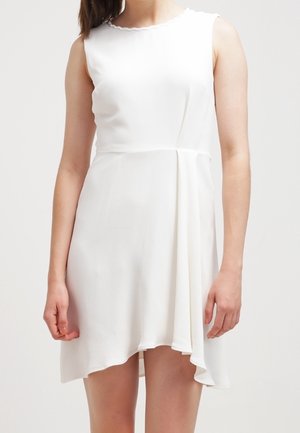 Frau trägt ein ärmelloses, weißes asymmetrisches Kleid mit einem dezenten gewellten Halsausschnitt und sanften Falten am Rock.