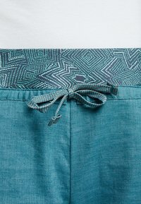 Teal broek met een gemusterde tailleband en een gestrikt koord in het midden voorop op een witte achtergrond.