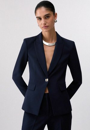 Donna che indossa un blazer blu navy su misura con un singolo bottone d'argento, pantaloni abbinati, collana girocollo d'argento e orecchini a cerchio d'argento.