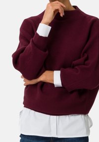 Suéter burdeos de canalé con cuello alto y mangas oversize, que presenta puños blancos. Llevado sobre una camisa blanca de botones.