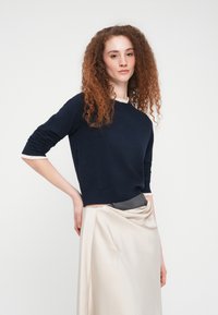 Γυναίκα με σγουρά κόκκινα μαλλιά, ντυμένη με μπλε σκούρο πουλόβερ και εκρού φούστα, που στέκεται με το ένα χέρι στη μέση της μπροστά σε απλό λευκό φόντο.