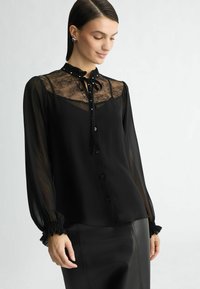 Mustarikk blouse, mis on valmistatud läbitavast kangast, pitsilise detailiga kaeluses, krael side ja nööbid ees. Pikad varrukad, millel on volangmühvid.